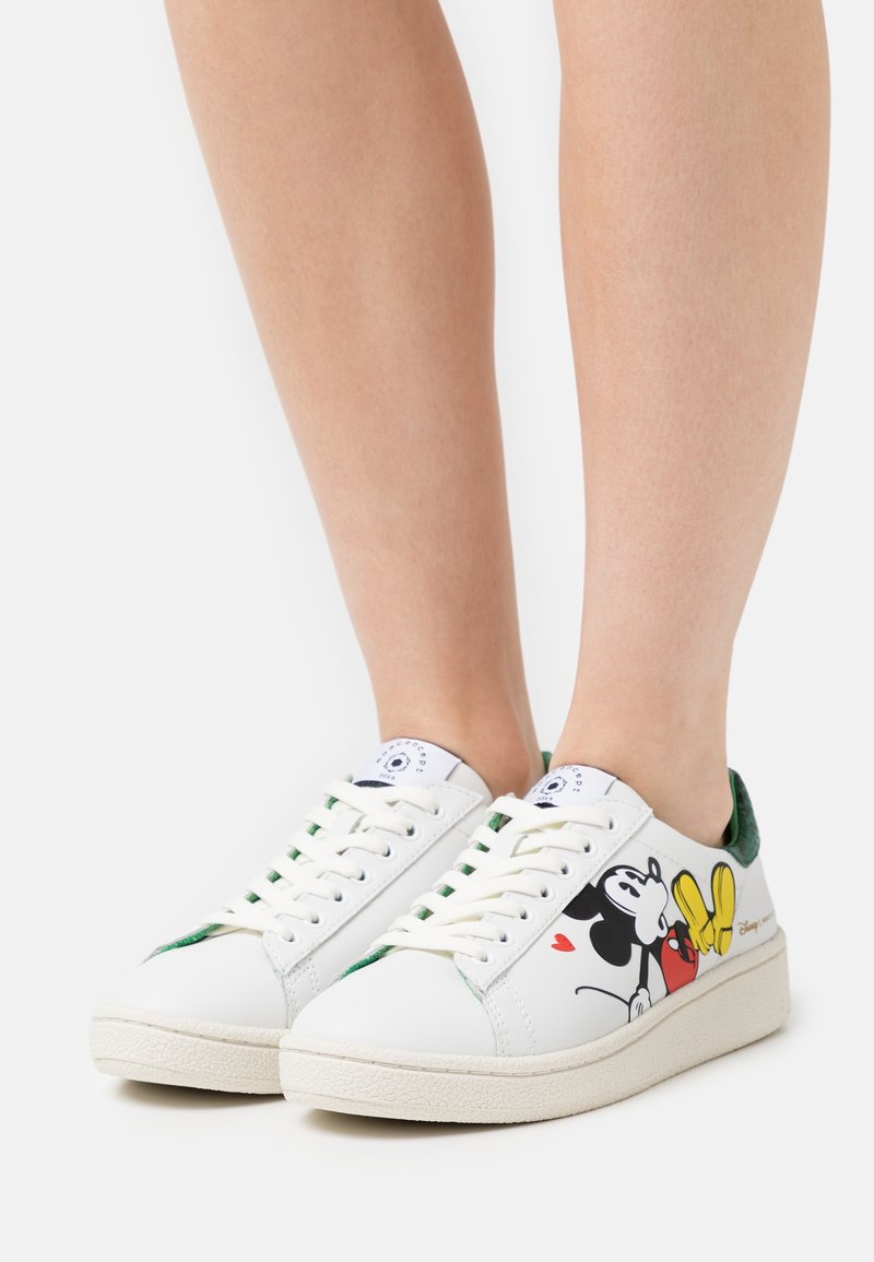 Vita sneakers med Mickey Mouse-mönster som bärs på fötterna, med gröna detaljer och krämfärgade sulor.