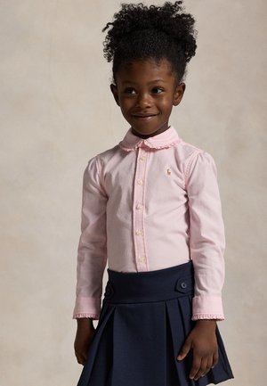 Jeune fille aux cheveux bouclés portant une chemise rose boutonnée et une jupe plissée bleu marine, souriant légèrement, debout devant un fond uni.