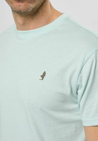 Miętowa bawełniana koszulka z okrągłym dekoltem i krótkimi rękawami. Posiada mały brązowy haftowany detal na lewej stronie klatki piersiowej.