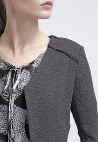Kvinna iklädd en texturerad mörkgrå jacka över en mönstrad svart och vit blus med knytning i halsen, som ser åt sidan mot en enfärgad bakgrund.