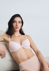 Lichtroze bh met schulpjes kant en een diamanten patroon, gecombineerd met een naakt hoge taille onderstuk, tegen een neutrale achtergrond.