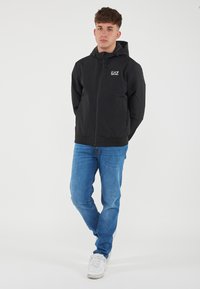 Black zip-up jacket with a hood, lightweight material, elastic cuffs, and hem, featuring a small white logo on the chest. Worn with blue jeans.