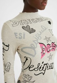 Haut à manches longues en rib en beige avec des textes et designs imprimés en noir et rose, y compris des cœurs, des fleurs et des étoiles. Encolure haute.