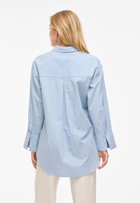 Gai zilgans kokvilnas krekls ar izkliedētu apkakli, garām piedurknēm un atpakaļ plecu detaļu, kam ir pogu aproces un noapaļots apakšmalas dizains.