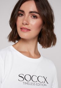 Camiseta de algodón blanca con la frase "SOCCX TIMLESS EDITION" en negrita y letras negras. Cuello redondo, mangas cortas, textura suave y diseño casual.