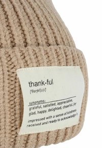 Beige knitted beanie featuring a textured rib design and a fabric label describing the word "thankful" with synonyms and definitions.