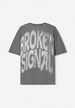 T-shirt gris surdimensionné avec un grand texte blanc usé et en lettres majuscules indiquant "SIGNAL COUPÉ" au dos. Manches courtes et col rond.