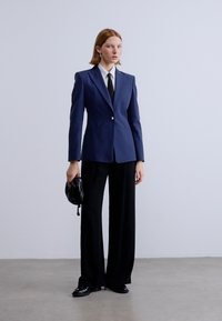 Ναυτικό σακάκι με κοφτερές πέτες, κλείσιμο με ένα κουμπί, σε συνδυασμό με μαύρο παντελόνι φαρδιάς γραμμής και μικρή μαύρη τσάντα, σε ουδέτερο φόντο.