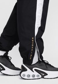 Pantalones deportivos negros con una franja lateral blanca y "SWOOSH AIR" en dorado, combinados con zapatillas en blanco y negro que cuentan con amortiguación de aire visible.
