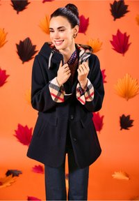 Marineblauer Wollmantel mit karierter Fleece-Innenausstattung und Bündchen, ausgestattet mit einer Kapuze, zwei Fronttaschen und einem Knopfverschluss, vor einem orangefarbenen Hintergrund.