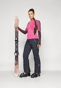 Top lungo aderente rosa e marrone con dettagli gialli, pantaloni da sci neri, scarponi da sci neri e uno sci con attacchi su uno sfondo neutro.