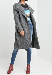 Cappotto grigio, texture, con design a davanti aperto, indossato sopra una maglietta grafica e jeans blu, abbinato a stivaletti alla caviglia rossi con stampa pitone.