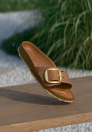Brune lædersandal med slide-design, guldbuckle, korkfodbed og tekstureret beige sål, løftet på en træoverflade mod grønne omgivelser.
