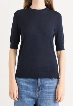 Femme portant un pull à manches courtes bleu marine foncé avec des poignets ajustés et un jean en denim bleu clair, debout devant un fond uni.