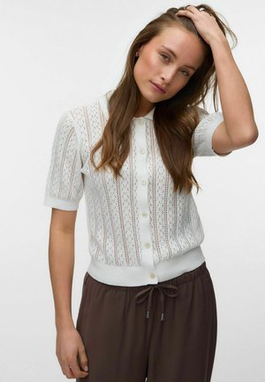 Ung kvinde med langt brunt hår iført en hvid kortærmet knapstrikket top og mørkebrune bukser med snørre, der rører ved sit hår.