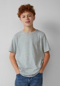 Ein rothaariger Junge trägt ein hellgraues T-Shirt und blaue Jeans mit den Händen in den Taschen und steht vor einem schlichten hellgrauen Hintergrund.