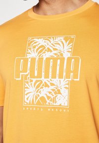 Žluté bavlněné tričko s bílým grafickým designem, který zobrazuje tropické listy a tučný text "PUMA" s nápisem "SPORTS RESORT" pod ním.