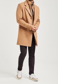 Tan ullkappa med slagkrage, enkel knappstängning och sidfickor, bärs över en brun polotröja och svarta byxor; kombinerad med vita sneakers.