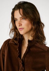 Blusa de pana marrón con cuello con volantes y lista de botones, con acentos metálicos en los hombros, textura suave y diseño ajustado.