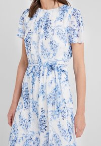 Blommig klänning i blått och vitt, med korta ärmar, en figurnära midja med knytning och ett strukturerat tyg med subtila mönster.