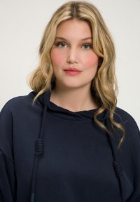 Marineblauwe hoodie met een relaxte pasvorm, voorzien van een trekkoordcapuchon en een gladde textuur; gedragen door een model met golvend blond haar en minimale make-up.