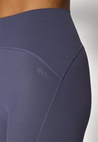 Sötétkék, magas derekú leggings, sima, rugalmas anyagból készült; lapos varrásokkal és az oldalon elhelyezett logóval rendelkezik.