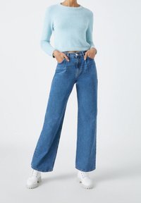 Suéter azul claro de felpa combinado con jeans de pierna ancha azul. La modelo lleva zapatos blancos con cordones. Los jeans tienen una cintura alta y bolsillos delanteros.