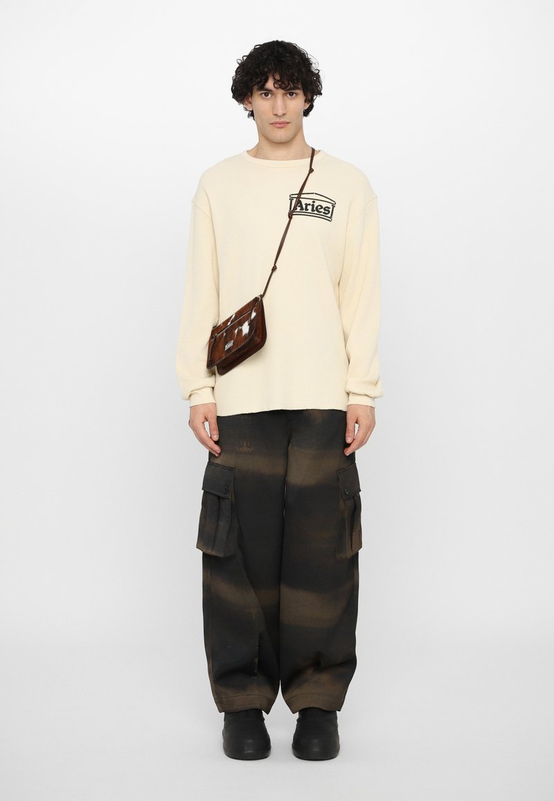 T-shirt à manches longues côtelé couleur crème avec un logo, associé à un pantalon cargo noir ample à motif camouflage. Porte un petit sac bandoulière marron.