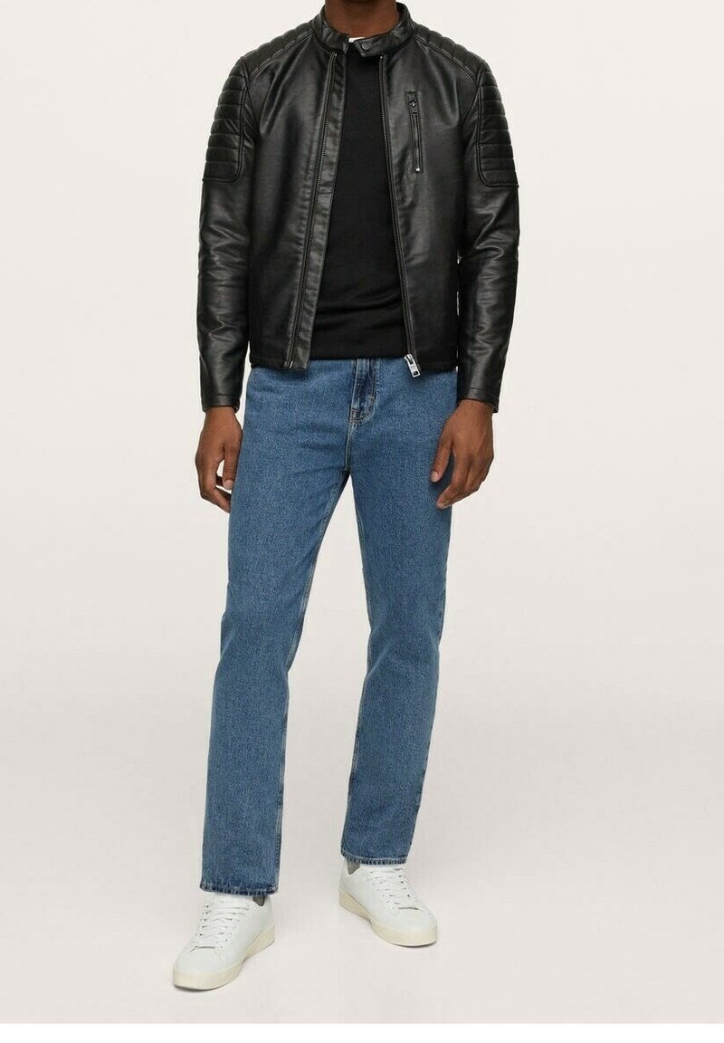Homme portant une veste en cuir noire, une chemise noire, un jean bleu et des baskets blanches, debout devant un fond clair uni.