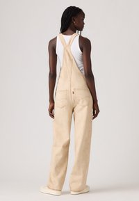 Beige denim overalls with adjustable shoulder straps, a loose fit, front pockets, and a branded back patch. Worn over a white tank top.