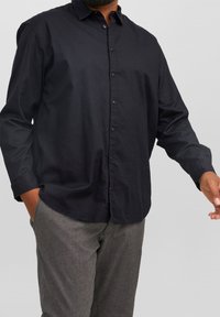 Camisa negra de botones hecha de tejido ligero, con cuello, mangas largas y un corte relajado, combinada con pantalones grises texturizados.