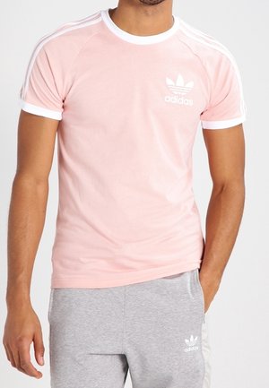 Homme portant un t-shirt Adidas rose clair avec bordure blanche et logo, associé à un pantalon de survêtement Adidas gris clair avec logo blanc et bande latérale.