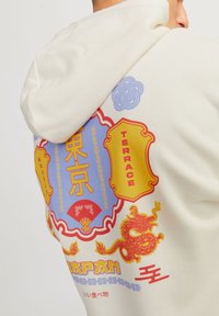 En person som bär en krämfärgad hoodie med färgglada grafiska motiv, inklusive en röd drake, tecken från Tokyo och orden "Terrace" och "Japan."