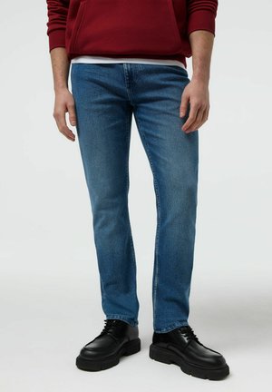 Mężczyzna ubrany w dopasowane niebieskie dżinsy, czarne masywne buty na sznurowadła oraz czerwony top z długim rękawem, stojący na jednolitym jasnym tle.