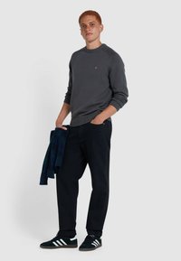 Grauer Strickpullover mit gerippten Bündchen, marineblaue Hose und schwarze Sneakers mit weißen Streifen. Hält ein dunkelgrünes karierter Hemd.