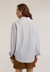 Blusa a righe azzurro chiaro con vestibilità rilassata, colletto e maniche lunghe. Presenta una leggera lucentezza e un orlo arrotondato. Indossata con pantaloni corti marroni.