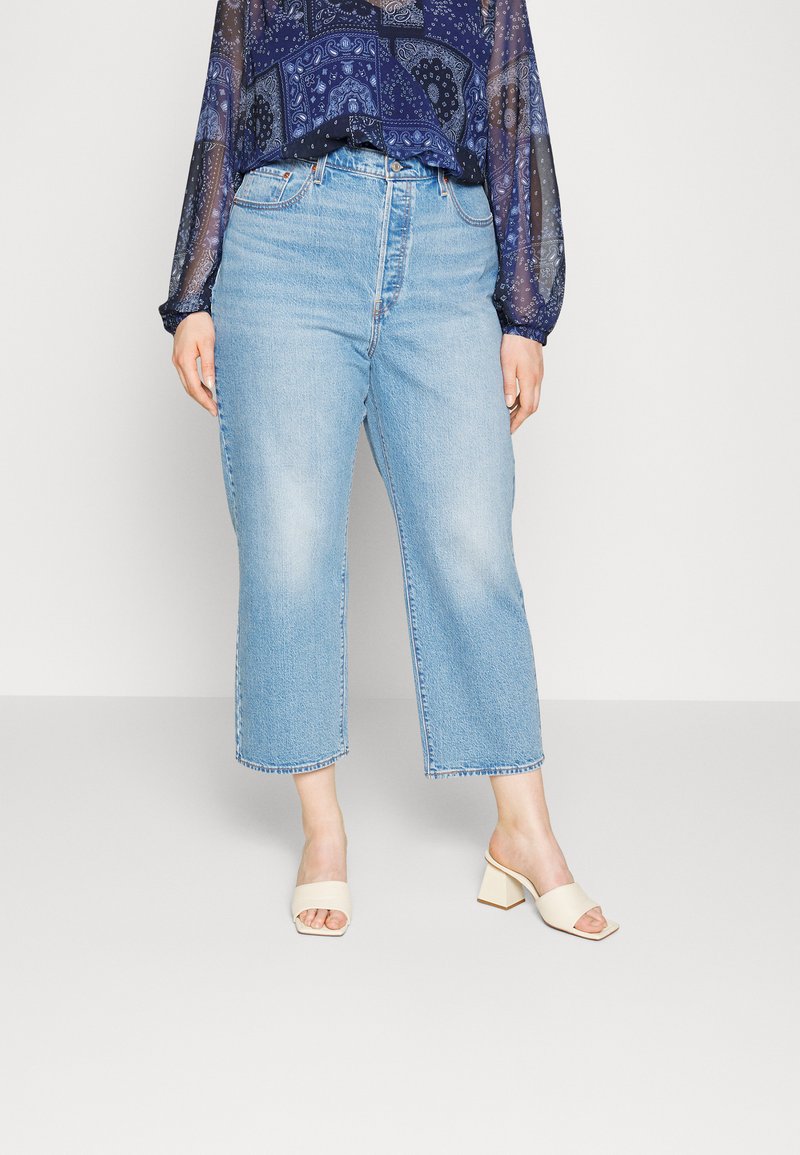 Helle blaue Denim-Jeans mit geradem, cropped Schnitt. Tragen mit einer dunkel gemusterten Bluse und beigen Blockabsatzsandalen.
