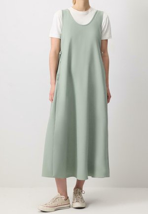 Langes, ärmelloses Kleid aus weichem grünem Stoff mit Rundhalsausschnitt, seitlichen Bändern und lockerem, fließendem Schnitt, kombiniert mit beigen Sneakern.