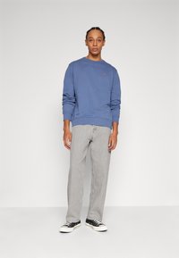 Blauwe sweatshirt van katoen, met een klein logo. Draagt bij lichte grijze, rechte jeans. Schoenen zijn zwart met witte zolen.
