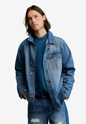 Junger Mann mit schulterlangem Haar, der eine abgestufte blaue Jeansjacke, passende abgenutzte Jeans und ein blaues Sweatshirt trägt, die Hände in den Taschen.