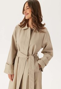 Beige trenchcoat med bälte i midjan, krage och långa ärmar. Har en rak passform och en slät textur.