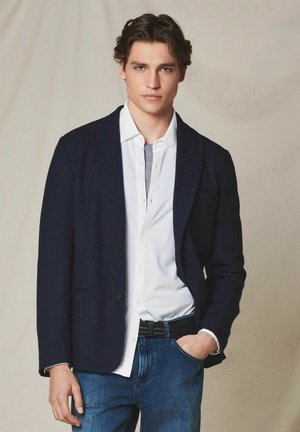 Junger Mann mit welligem braunem Haar trägt einen dunklen Blazer, ein weißes Hemd über einem grauen Unterhemd und blaue Jeans, mit einer Hand in der Tasche.