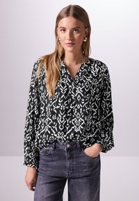 Schwarz-weiß gemusterte Bluse mit V-Ausschnitt, langen Ärmeln und gerafften Details an den Manschetten, kombiniert mit hochgeschnittenen grauen Jeans.