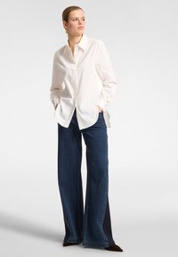 Weißes Hemd mit langen Ärmeln und goldenen Knöpfen an den Manschetten, kombiniert mit dunkelblauen weiten Jeans. Glatte Texturen, lässige Passform.