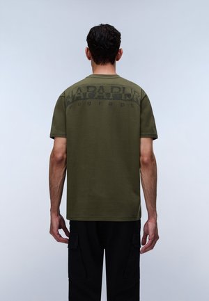 Homme de dos portant un T-shirt vert olive à manches courtes avec un texte noir sur le haut du dos et un pantalon noir avec poches latérales.