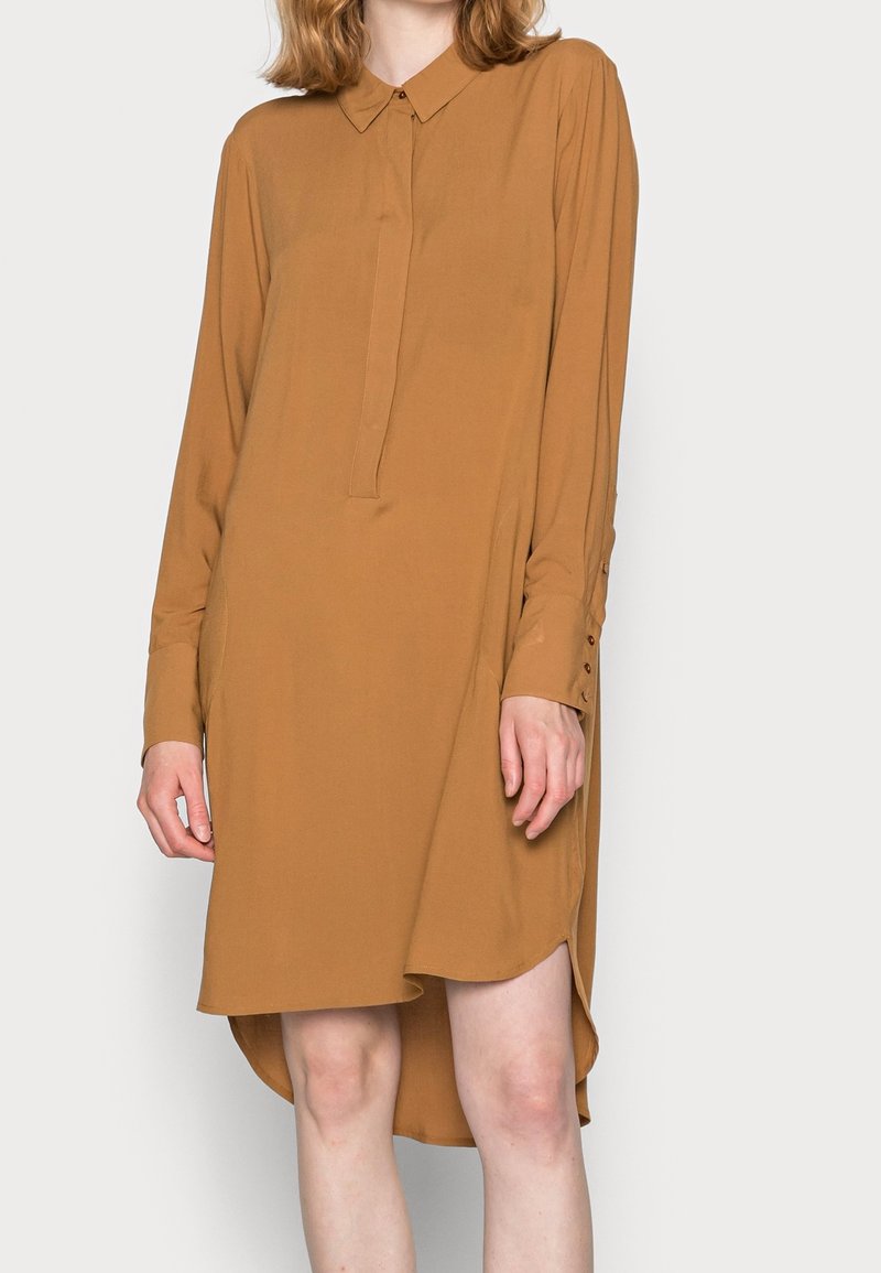 Robe chemise à manches longues en tissu brun uni, avec un col à boutons, une patte de boutonnage à l'avant et des fentes sur les côtés pour le mouvement.