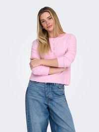 Ženska z dolgimi blond lasmi, oblečena v roza pulover z gumbi in modre kavbojke, stoji z prepognjenimi rokami pred enobarvnim ozadjem.