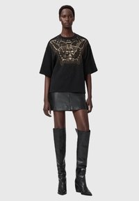 Camiseta negra oversize con diseño gráfico dorado, combinada con una minifalda de cuero negra y botas altas negras de tacón.
