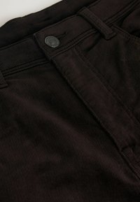 Sorte kordfløjlstof med en ribbet tekstur, præsenteret med en frontknaplukning og bæltestropper i et nærbillede.