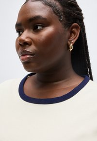 Κοντινό πλάνο γυναίκας με πλεγμένα μαλλιά που φοράει χρυσά και μαργαριταρένια σκουλαρίκια και μια κρεμ μπλούζα με στρογγυλή λαιμόκοψη σε ναυτικό μπλε χρώμα.